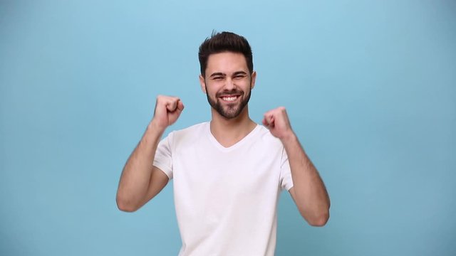 Handsome unshaven bearded young guy 20s wearing white t-shirt isolated on pastel blue background in studio. People sincere emotions lifestyle concept. Walking showing thumbs up pointing fingers camera