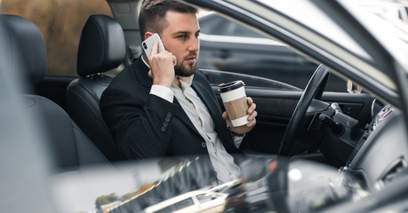 Handsome caucasian businessman driver inside in vehicle  with coffee and gadget and check mail and talking with partners