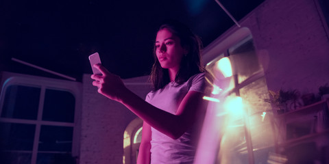 Calm. Cinematic portrait of handsome stylish woman in neon lighted interior. Toned like cinema effects in purple-blue. Caucasian female model using smartphone in colorful lights indoors. Flyer.