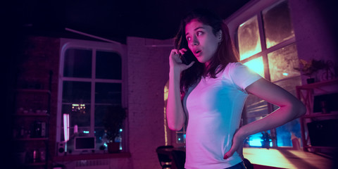 Talking on phone. Cinematic portrait of handsome stylish woman in neon lighted interior. Toned like cinema effects in purple-blue. Caucasian model using smartphone in colorful lights indoors. Flyer.