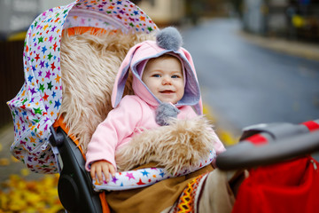 Cute little beautiful baby girl sitting in the pram or stroller on autumn day. Happy smiling child in warm clothes, fashion stylish pink baby coat with bunny ears. Baby going on a walk with parents.