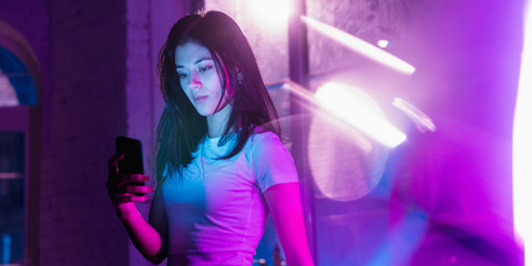 Serfing attented. Cinematic portrait of handsome stylish woman in neon lighted interior. Toned like cinema effects in purple-blue. Caucasian model using smartphone in colorful lights indoors. Flyer.