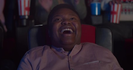 Close up of happy young man face watching comedy film in cinema. Millennial african guy sitting in chair and laughing in movie theatre. Concept of entertainment. Positive emotions.