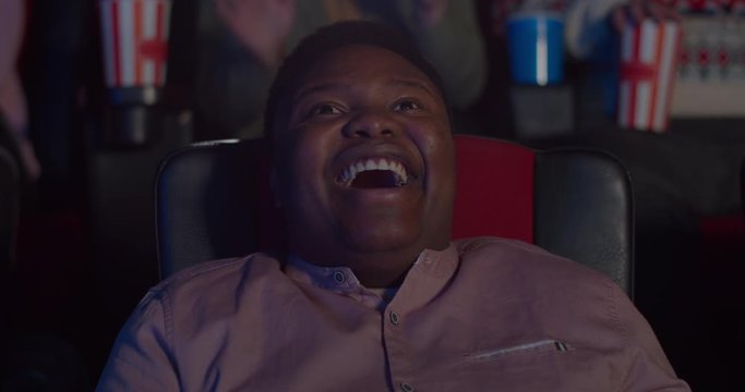 Close up of happy young man face watching comedy film in cinema. Millennial african guy sitting in chair and laughing in movie theatre. Concept of entertainment. Positive emotions.