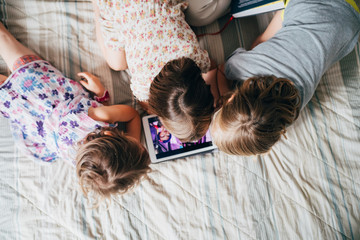 Overhead view of three little sisters indoor at home using tablet lying in bed