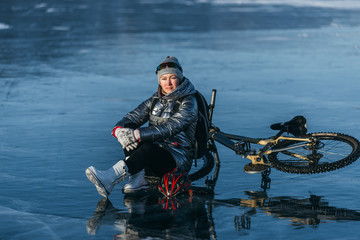 Woman sitting near her bike on ice. The girl cyclist stopped to rest. She sits on the wheel and enjoys the beautiful view of the sunset. The traveler is ride a cycle. Female is riding bike on the ice.