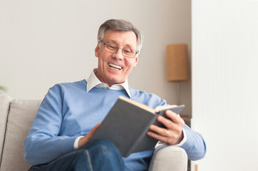Senior Man Reading Book Sitting On Sofa At Home