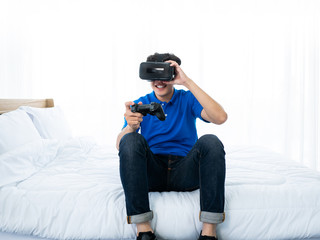 Obraz premium Asian young man enjoy and playing virtual reality goggles simulation digital technology device to connect game online with joystick controller gadget equipment hobbies and leisure at white bedroom