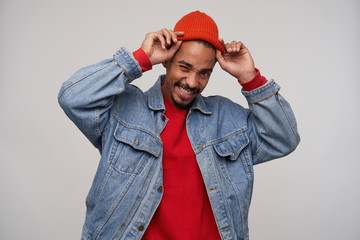 Cheerful young pretty dark skinned brunette male with beard holding his red cap with raised hands while standing over white background, being in nice mood and smiling happily