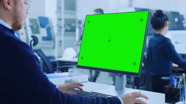 Over the Shoulder: Heavy Industry Engineer Working on Personal Computer, Display is Green Mock-up Screen or Chroma Key. Industrial Factory with High-Tech CNC Machinery