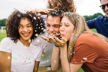 Group of four friends having fun at the park blowing confetti - Millennials playing together at...