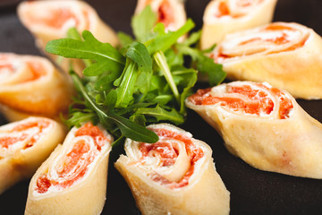 Rolled pancakes (crepes, blini) with smoked salmon and cream cheese served with arugula on black plate.