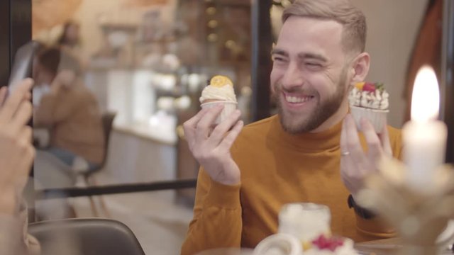 Medium shot of young woman making photos of Caucasian handsome bearded man sitting at cafe table with cupcakes in his hands and having fun