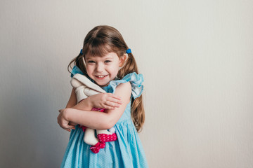 Pretty little girl in blue dress smiling at camera and holds her toy rabbit
