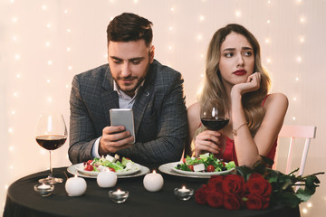 Boring Date. Man using smartphone in restaurant, ignoring his sad girlfriend