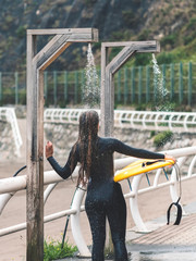 back view of a surfer girl in a wetsuit taking a shower on the beach with her board in her hand...