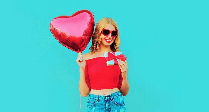 portrait happy smiling young woman holding gift box, red heart shaped air balloon on colorful blue background