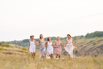 The company of cheerful female friends have a great time together on a picnic in a picturesque place overlooking the green hills. Girls in white dresses dancing in the field