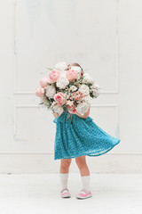 Cute little girl child in a summer dress holds a large bouquet of flowers near a white wall. Studio...