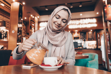 Muslim woman pours tea from a teapot into a cup while sitting at a table in a cafe