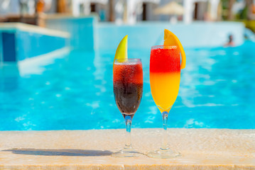 Two cocktails with natural juice and ice at the edge of the swimming pool. Orange cocktail with an orange slice and red with a melon slice with backdrop of vibrant pool water.