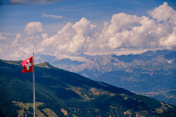 Swiss flag waving high up in the Alps near alpine village Saint-luc in the Val d' Anniviers in summer on a cloudy day. Valais, Switzerland