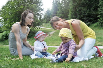 Women sitting with little baby girls on grass in the park. Two friend women enjoying a beautiful sunny day with their little daughters. Concept of motherhood, friendship and summer vacations.