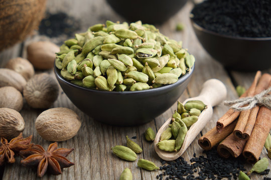 Green Cardamom Pods In Black Ceramic Bowl. Aromatic Spices: Anise, Gloves, Black Cumin Seeds, Nutmegs, Cinnamon Sticks, Turmeric. Ingredients For Healthy Cooking. Ayurvedic Treatments.