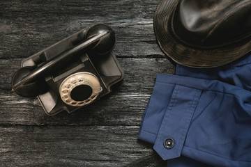 Old black rotary phone, coat and leather hat on black wooden desk background.