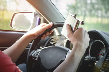 Drunk driver is driving a car on the road and drinking a strong alcohol from a flask close up.