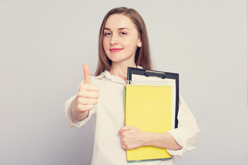 Happy woman student with notebooks shows thumb up. Concept of education. Portrait, grey background, copy space, slogan, toned