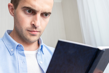 Serious guy reading a book sitting on sofa at home, portrait, close up, toned