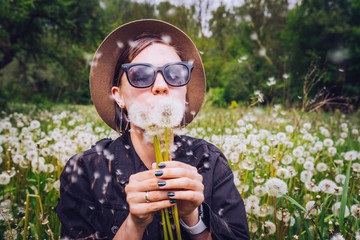 Happy woman blowing on dandelion in park. Girl in hat and sunglasses. Wishing, joy concept