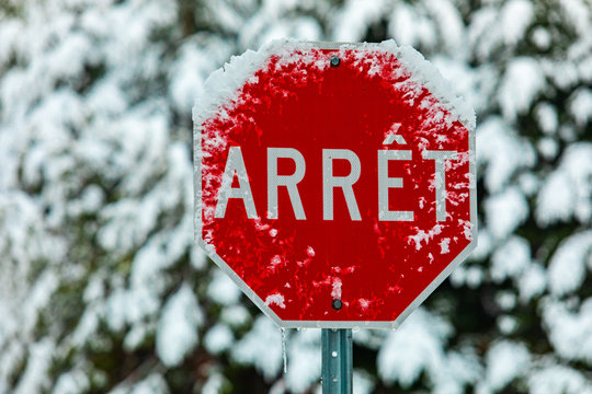 A close up selective focus shot of a traffic sign saying Arret, French for Stop, seen on a street in Quebec during winter, covered in snow with blurry trees
