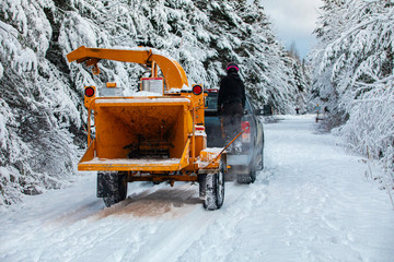 A wide view of an arborist standing on the back of a truck pulling a mobile tree chipper on a rural road during winter, heavy snow causes disruption