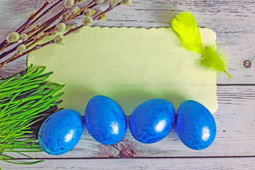Easter background with easter eggs. Yellow sheet of paper for text, four blue easter eggs, willow branches, feathers. on a wooden light background. Top view, save the space. Happy Easter Greeting Card