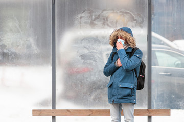 Sick man with a hood alone on public transport stop, coughing, wearing protective facial mask against transmissible infectious diseases and as protection against the flu or coronavirus in public place