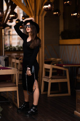 Fototapeta na wymiar Photo of brunette lady posing on wooden background in cafe.Fashion style portrait. Girl wearing dark casual dress, sunglasses and dark hat .Fashion concept.