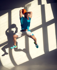 Young athletic basketball player jumping with ball in front of wall with shadows from window