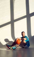 Young athletic man, basketball player holding ball with one hand and sitting near wall with shadows...