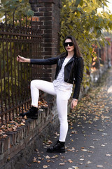 Outdoor fashion portrait of stylish young woman having fun, emotional face , laughing, Urban city street style.Girl wearing white pants,t-shirt, black leather jacket , sunglasses and dark hat Fashion.