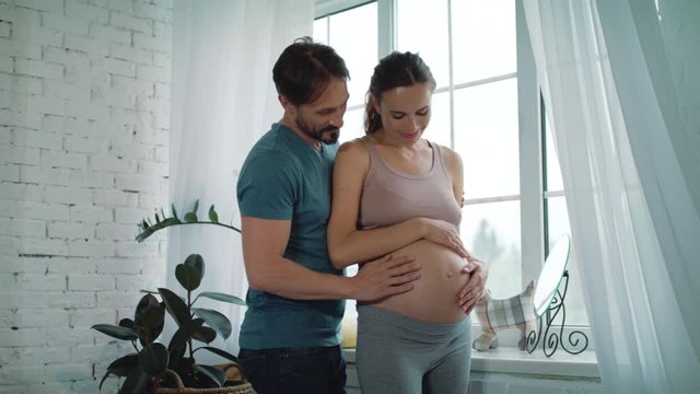 Happy husband holding belly og pregnant wife at home. Pregnant couple hugging