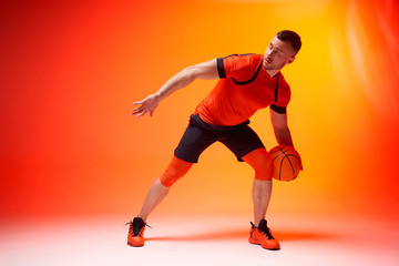 Fototapeta na wymiar Young athletic basketball player in attack position holding ball with one hand on orange and red background