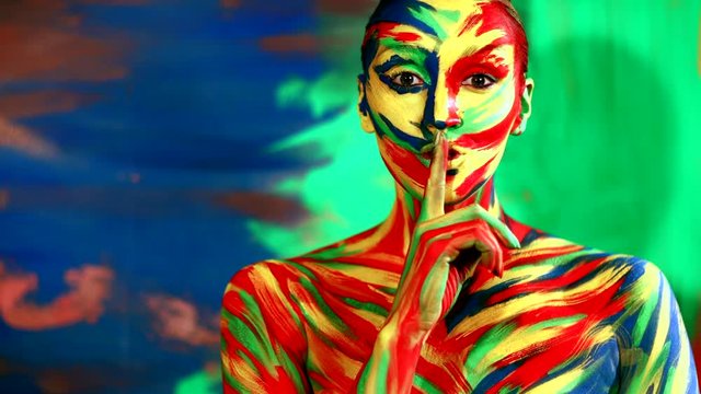 Color face art and body paint on woman showing silence sign the finger near lips. Abstract portrait of the bright beautiful girl with colorful make-up and bodyart.