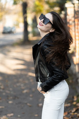 Outdoor fashion portrait of stylish young woman having fun, emotional face , laughing, Urban city street style.Girl wearing white pants,t-shirt, black leather jacket , sunglasses and dark hat Fashion.