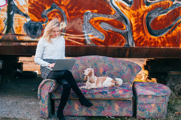 Cheerful caucasian young freelancer girl sitting on old couch outdoor over rusty graffiti wall on background and using on laptop with her lovely cocker spaniel dog lying close to her.