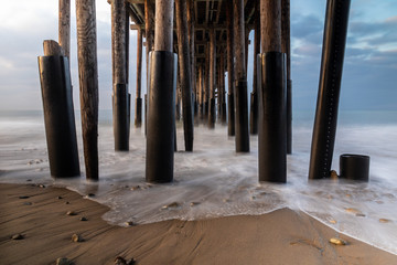 Closeup of pillars, Ventura Pier, Ventura, California. Early light from sunrise visible from left.  sand and rocks in foreground; smooth incoming wave reaching shore; ocean, sky beyond. 