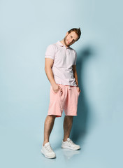 Young smiling handsome man in pink shorts and white polo shirt standing and looking at camera over light blue wall