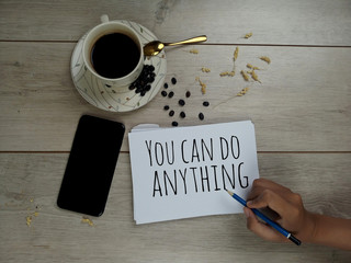 Inspirational quote - You can do anything. With a cup of hot black morning coffee, roasted coffee beans, hand phone, and a young woman hand with pencil writing on white paper note book on wooden table