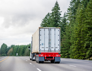 Big rig semi truck transporting cargo in long container mounted on a flat bed semi trailer running on the wide road with green trees on the side